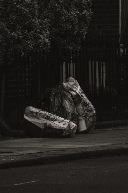 The image depicts two large, tightly packed bags of mixed rubbish placed on a city pavement near a black metal fence and a brick wall. The bags are made of clear and white plastic, filled with various