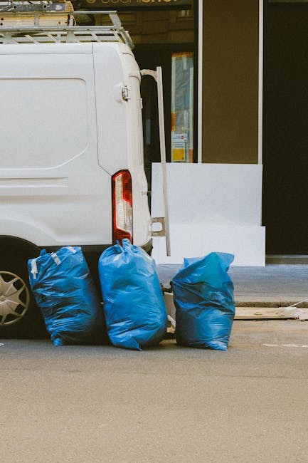 The image depicts two large, tightly packed bags of mixed rubbish placed on a city pavement near a black metal fence and a brick wall. The bags are made of clear and white plastic, filled with various